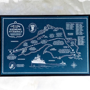 May include: A framed navy blue map print detailing the route and lore of the Edmund Fitzgerald. The map features illustrations of the ship, portraits, and text including the ship's name and locations like Duluth and Whitefish Point.