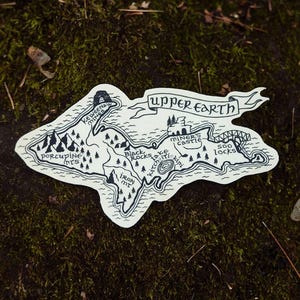 May include: A black and white sticker with a map of a region, featuring text that reads "Upper Earth." The map includes illustrations of mountains, trees, and bodies of water, with place names like "Porcupine Mts" and "Soo Locks."