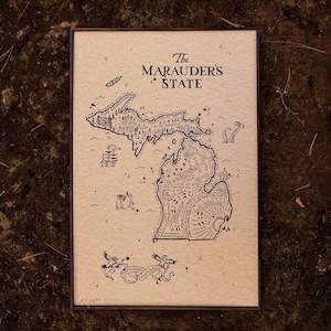 May include: A framed art print featuring a map of Michigan with intricate black line art. The text "The Marauder's State" is at the top. The map includes illustrations of a ship, sea creatures, and other whimsical elements.
