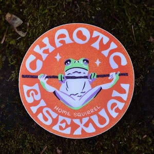 May include: A round sticker with an orange background and white text that reads "CHAOTIC BISEXUAL HOME SQUIRREL." A cartoon frog is centered, holding a bar. The frog is green, white, and purple. Two white stars are on either side of the frog.