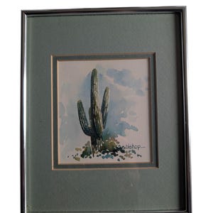 May include: Framed watercolor painting of a saguaro cactus. The artwork features a green cactus with three arms, set against a light blue sky with clouds. The painting is signed "bishop" and framed in a silver metal frame.