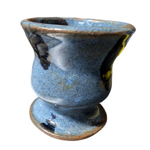 Può includere: Un piccolo vaso in ceramica fatto a mano con una glassa blu. Il vaso ha un corpo arrotondato, un bordo svasato e una base a piede. Il bordo e la base hanno un bordo marrone. Il vaso è alto circa 10 cm.