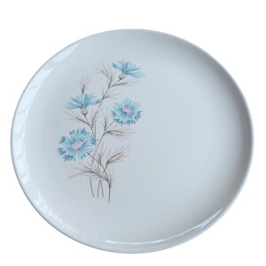 May include: A white ceramic plate with a floral design. The plate features a cluster of blue flowers with pink centers and gray-green leaves. The flowers are arranged on a stem, and the plate has a smooth, glossy finish.