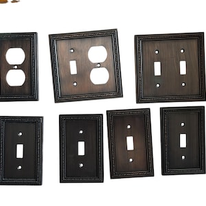 May include: A collection of bronze-colored electrical switch plates and outlet covers. The plates have a rectangular shape with a beaded border. Various configurations include single and double switch plates, and outlet covers.