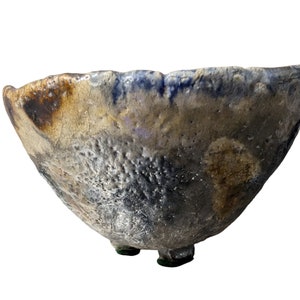 Può includere: Una ciotola in ceramica fatta a mano con una superficie unica e strutturata. La ciotola presenta un mix di colori, tra cui marrone, blu e grigio, con un bordo leggermente irregolare. Poggia su tre piccoli piedi, mostrando un'estetica rustica e artigianale.