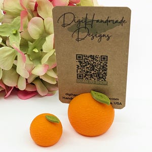 May include: Two orange-colored, textured, round objects with green leaf accents, resembling oranges, are displayed. A brown card with the text "Digit Handmade Designs" and a QR code is behind the oranges. Pink and green flowers are in the background.