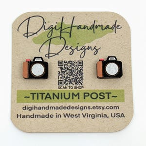 Puede incluir: Un par de pendientes de madera con forma de cámara con un acento rojo en la lente. Los pendientes están en una tarjeta marrón con el texto "DigiHandmade Designs" y "SCAN TO SHOP" impreso en ella. El texto "TITANIUM POST" está impreso en verde debajo del código QR. El texto "digihandmadedesigns.etsy.com" y "Handmade in West Virginia, USA" está impreso debajo del texto "TITANIUM POST".