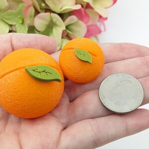 May include: Two orange-colored, round decorative objects, each with a green leaf accent, are displayed. A U.S. quarter is next to the objects for size comparison. The objects are likely miniature food replicas or decorative items.