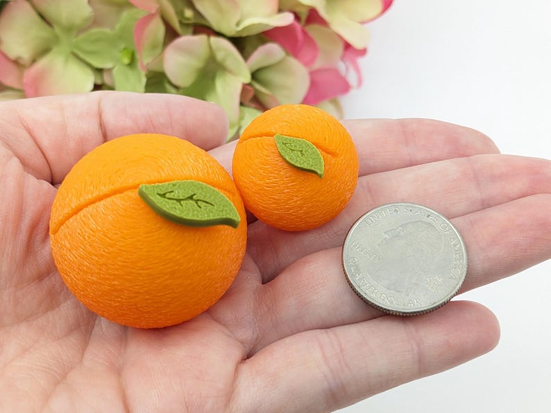 May include: Two orange-colored, round decorative objects, each with a green leaf accent, are displayed. A U.S. quarter is next to the objects for size comparison. The objects are likely miniature food replicas or decorative items.