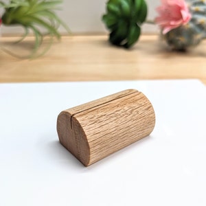 May include: A wooden, cylindrical place card holder with a slot in the center. The holder is light brown and has a natural wood grain.
