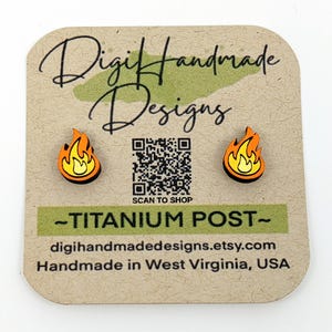 May include: A pair of wooden earrings with a flame design. The earrings are orange and yellow with a black outline. The earrings are on a brown card with the text "Digi Handmade Designs" and a QR code. The text "SCAN TO SHOP" is below the QR code. The text "-TITANIUM POST-" is below the QR code. The text "digihandmadedesigns.etsy.com" is below the text "-TITANIUM POST-". The text "Handmade in West Virginia, USA" is below the text "digihandmadedesigns.etsy.com".