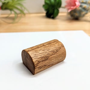 May include: A small, wooden, brown, cylindrical table number holder with a slot for a card. The holder is made of natural wood with a rustic finish.
