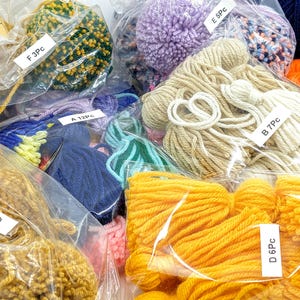 May include: Assortment of colorful yarn and pom poms in clear plastic bags. Each bag is labeled with a letter and number indicating the quantity of yarn or pom poms. The labels read: A 12PC, B 7PC, C 5PC, D 6PC, E 5PC, F 3PC.