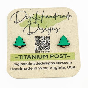 May include: A pair of green wooden Christmas tree earrings with titanium posts. The earrings are on a card that says "Digi Handmade Designs" and has a QR code that says "Scan to Shop". The card also says "Titanium Post" and "digihandmadedesigns.etsy.com". The card also says "Handmade in West Virginia, USA".