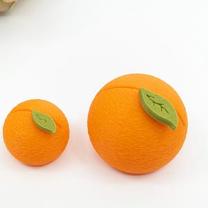 May include: Two orange-colored, spherical objects resembling oranges, each with a green leaf attached. The larger orange is approximately 3 inches in diameter, while the smaller one is about 2 inches across. The objects are set against a white background.