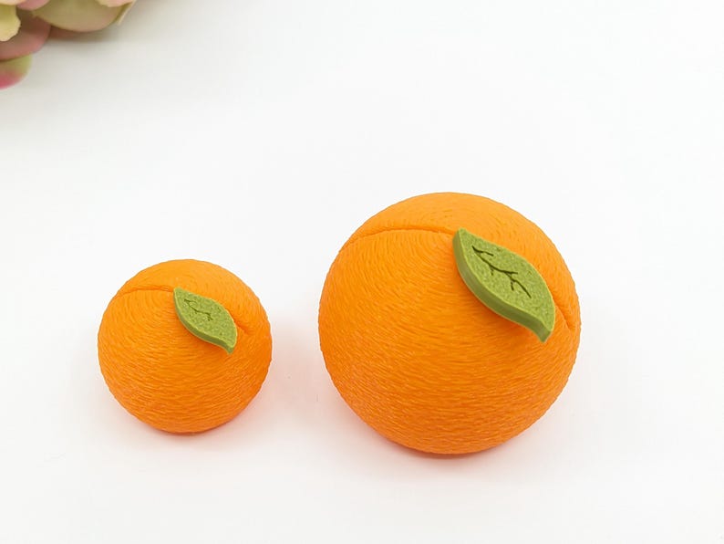 May include: Two orange-colored, spherical objects resembling oranges, each with a green leaf attached. The larger orange is approximately 3 inches in diameter, while the smaller one is about 2 inches across. The objects are set against a white background.