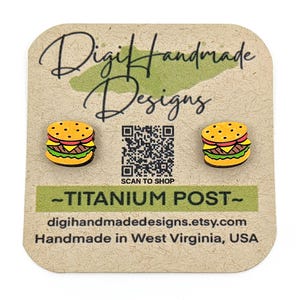 May include: A pair of wooden stud earrings featuring a cartoon burger design. The earrings are on a brown card with the text "Digi Handmade Designs" and "SCAN TO SHOP". The card also includes the text "TITANIUM POST" and "digihandmadedesigns.etsy.com Handmade in West Virginia, USA".