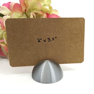 May include: A brown rectangular card, measuring 2" x 3.5", is held by a silver, dome-shaped holder. The card is in front of a bouquet of pink and yellow flowers, set against a white background.
