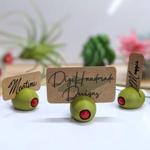 May include: Three olive-shaped place card holders with brown paper cards. The cards have handwritten text: "Martini", "Digi Handmade Designs", and "Maggie".