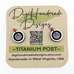 May include: A pair of wooden earrings featuring a target with an arrow in the center. The earrings are on a card that says "Digi Handmade Designs" and "Titanium Post".