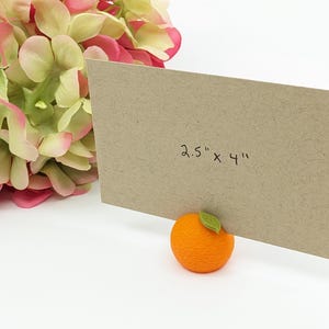 May include: A brown card, measuring 2.5" x 4", is propped up by an orange-shaped holder with a green leaf. Pink and green flowers are in the background. The card is made of textured paper.