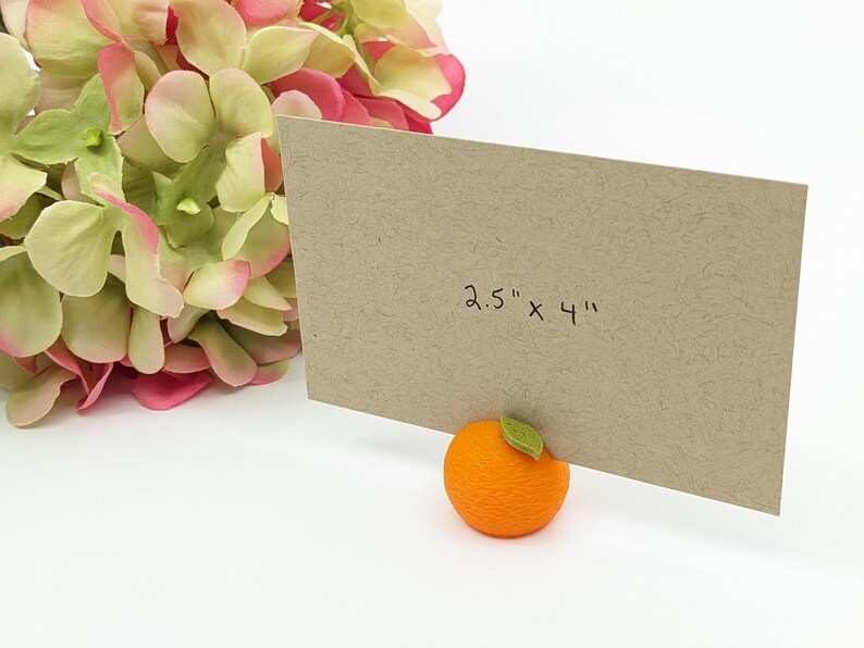 May include: A brown card, measuring 2.5" x 4", is propped up by an orange-shaped holder with a green leaf. Pink and green flowers are in the background. The card is made of textured paper.