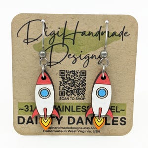 May include: A pair of rocket-shaped earrings with red, white, and blue details. The earrings are hanging on a brown card with the text "Digit Handmade Designs" and "Daisy Danles".
