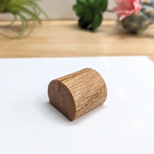 May include: A wooden place card holder with a natural wood finish. The holder is shaped like a half-cylinder with a slot in the center for holding a card.