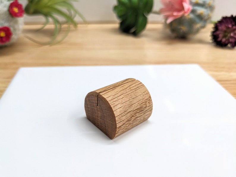 May include: A wooden place card holder with a natural wood finish. The holder is shaped like a half-cylinder with a slot in the center for holding a card.
