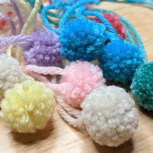 May include: A collection of colorful yarn pom-poms in various shades of blue, pink, yellow, green, and purple. The pom-poms are made from soft, fluffy yarn and are perfect for adding a touch of whimsy to any craft project.