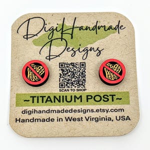 May include: A pair of wooden earrings with a red circle and a black no peanut symbol. The earrings are on a brown card with the text "Digi Handmade Designs" and "SCAN TO SHOP". The text "TITANIUM POST" is also on the card.