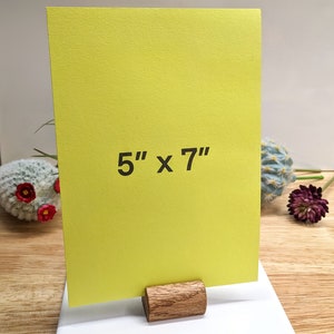 May include: A bright yellow card with the text "5" x "7" printed on it. The card is standing on a wooden easel.