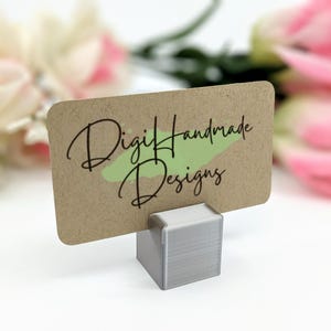 May include: A kraft paper card with rounded corners, featuring the text "Digi Handmade Designs" in black script over a green paint stroke. The card is held by a small, gray, cube-shaped holder.