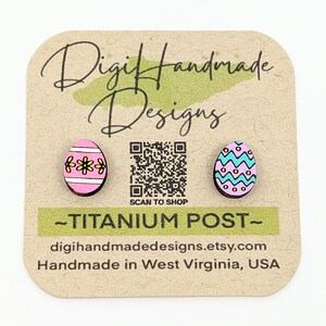 May include: Wooden Easter egg stud earrings. One earring is pink with a yellow flower design, the other is blue, purple, and yellow. The earrings are on a brown card with the text "Digit Handmade Designs" and "-TITANIUM POST-".