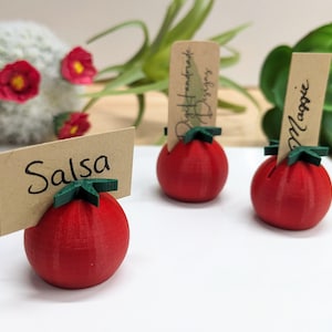 May include: Three red tomato-shaped place card holders with green leafy tops. One holder has a card that reads "Salsa", another reads "Right handmade Designs", and the third reads "Maggie". The holders are on a white surface.