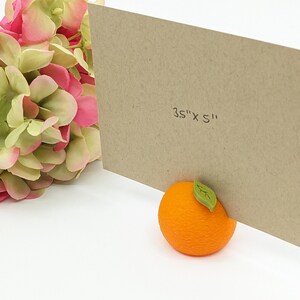 May include: A kraft paper card, measuring 3.5" x 5", is propped up by an orange-shaped object with a green leaf. Pink and green hydrangea flowers are in the background. The card is a neutral color, and the orange is bright.