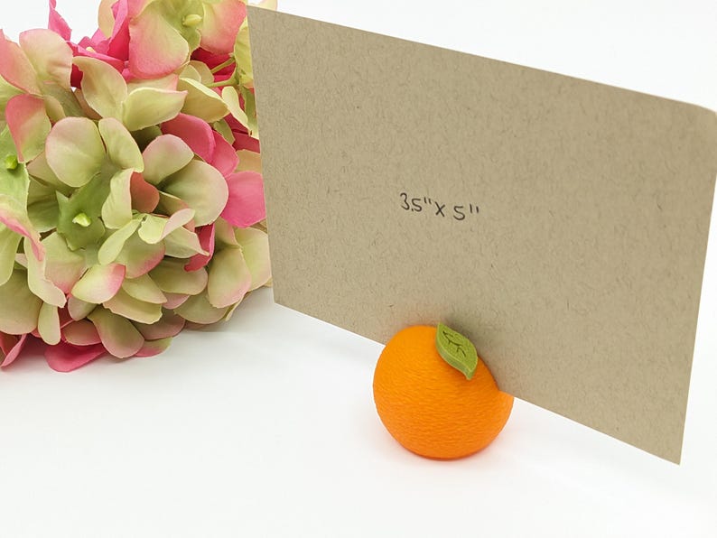 May include: A kraft paper card, measuring 3.5" x 5", is propped up by an orange-shaped object with a green leaf. Pink and green hydrangea flowers are in the background. The card is a neutral color, and the orange is bright.