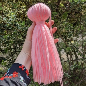 May include: A pink yarn tassel with a round top. The tassel is made of soft, fluffy yarn and has a long, flowing fringe.