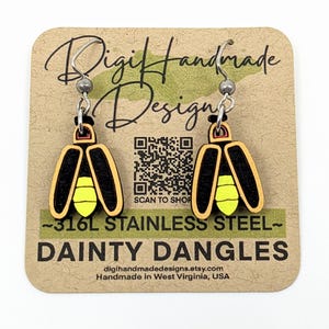 May include: Firefly-shaped earrings with black, yellow, and tan details. The earrings are on a tan card with the text "Digi Handmade Designs" and "Dainty Dangles". The earrings have stainless steel hooks.