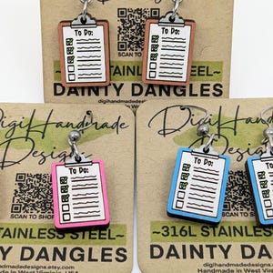 May include: Dainty dangle earrings featuring a miniature to-do list design. The earrings have a brown, pink, and blue border with a white background and black text that reads "To Do:". The earrings are made of stainless steel.