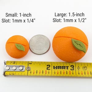 May include: Two orange-colored, spherical objects with green leaf accents, one 1-inch and the other 1.5-inch, are displayed next to a US quarter and a yellow measuring tape. Text indicates slot sizes.