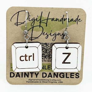 May include: A pair of dangle earrings featuring white square keyboard key designs with the words "ctrl" and the letter "Z" in brown. The earrings are attached to silver-colored hooks and are displayed on a brown card with the text "DAINTY DANGLES".