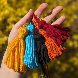 May include: Five colorful yarn tassels in shades of yellow, blue, orange, red, and black. The tassels are tied together and held in a hand.