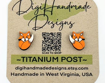 May include: A pair of orange fox stud earrings with titanium posts. The earrings are displayed on a card with the text "Digi Handmade Designs", a QR code, and the text "SCAN TO SHOP", "TITANIUM POST", "digihandmadedesigns.etsy.com", and "Handmade in West Virginia, USA".