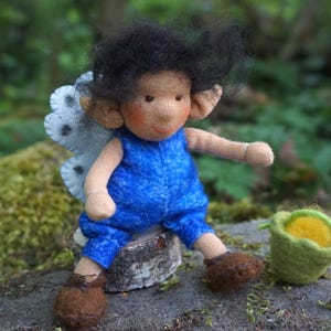 Könnte beinhalten: Eine handgefertigte Filzpuppe mit dunklem, zerzaustem Haar, spitzen Ohren und rosigen Wangen, bekleidet mit einem leuchtend blauen Strampler. Die Puppe hat weiße Filzflügel mit schwarzen Punkten und einen kleinen grünen Filzeimer mit gelbem Inhalt.
