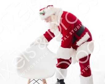 May include: Santa Claus in a red suit with white trim and a matching hat, bending over a white chair with wooden legs. He wears black boots and a black belt. The scene is set against a white background, evoking a festive holiday theme.