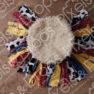 Puede incluir: Un accesorio redondo y esponjoso de color crema para fotografía de recién nacidos, con una franja de tela alrededor del borde. La franja está hecha de varias telas, incluyendo estampado de bandana rojo y blanco, mezclilla azul, amarillo y estampado de vaca blanco y negro.