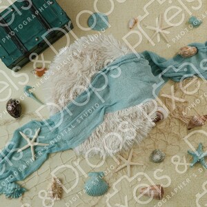 May include: A beach-themed newborn photography prop setup. Includes a teal chest, starfish, seashells, a blue wrap, and a fluffy white blanket, all arranged on a textured, sand-colored surface. The scene is decorated with a fishing net.
