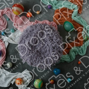 May include: A gray fuzzy round pouf sits on a gray background surrounded by pink, white, and blue fabric scraps and colorful balls.