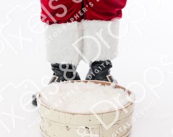 May include: A festive scene featuring Santa Claus's legs in red pants with white fur trim and black boots. A distressed, off-white wooden tub filled with white material sits in front of him, creating a holiday theme.